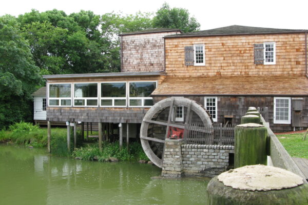 A rustic watermill beside a calm pond surrounded by greenery.